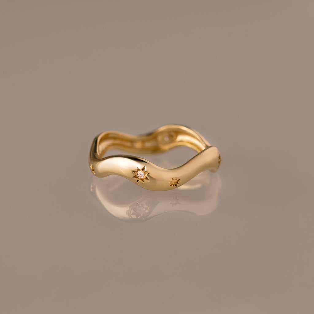 The Wavy Diamond Celestial Ring, featuring star-shaped engravings, is photographed on a reflective beige surface.