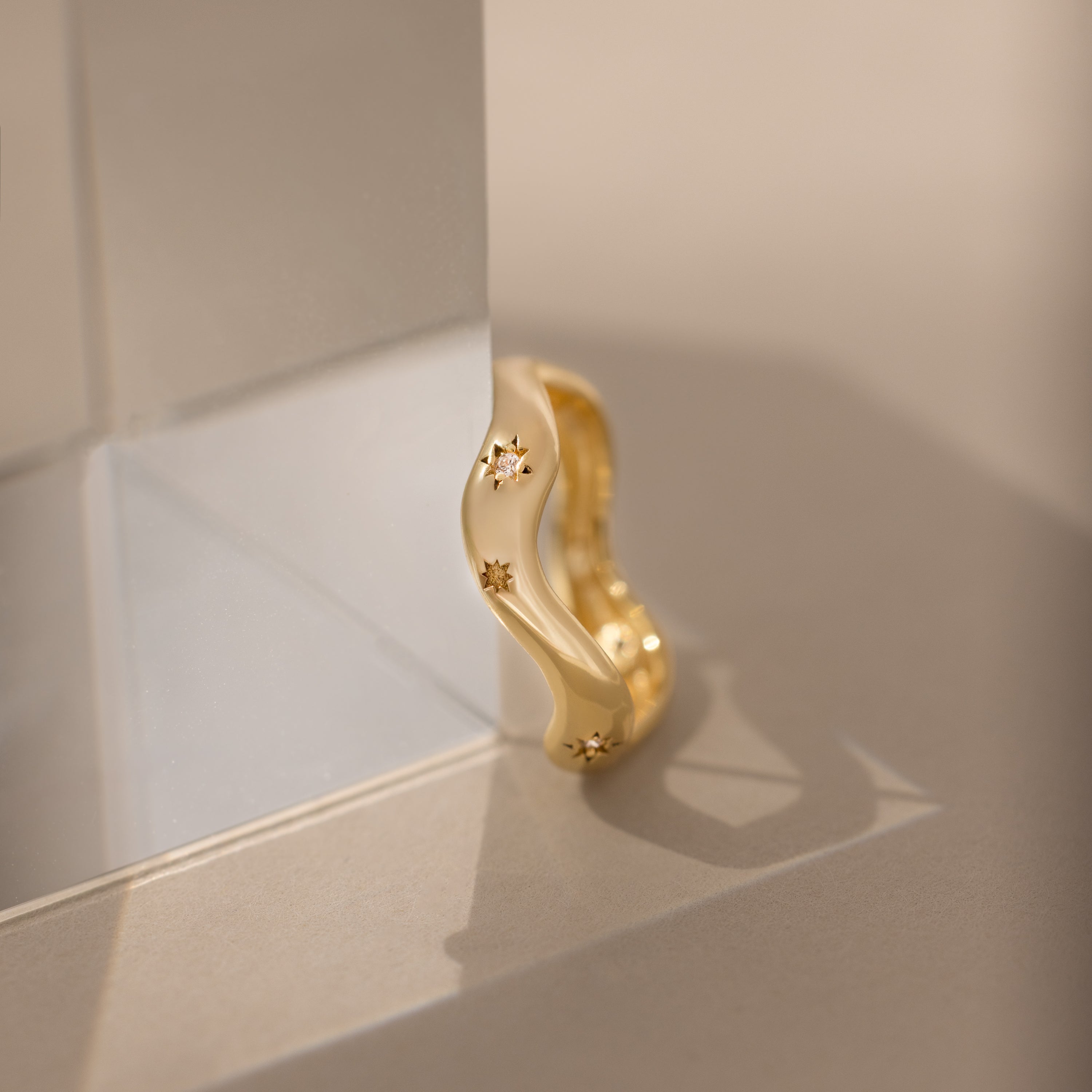 The Wavy Diamond Celestial Ring features small star-shaped engravings and is pictured resting against a clear glass block on a beige surface.
