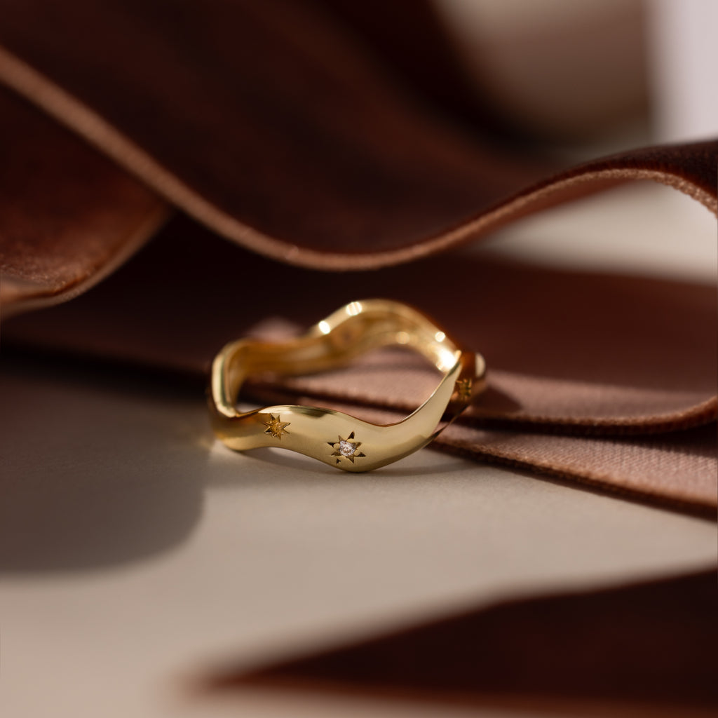 The Wavy Diamond Celestial Ring, adorned with star engravings, rests on soft brown fabric under warm light, capturing the enchanting sparkle of celestial diamonds.