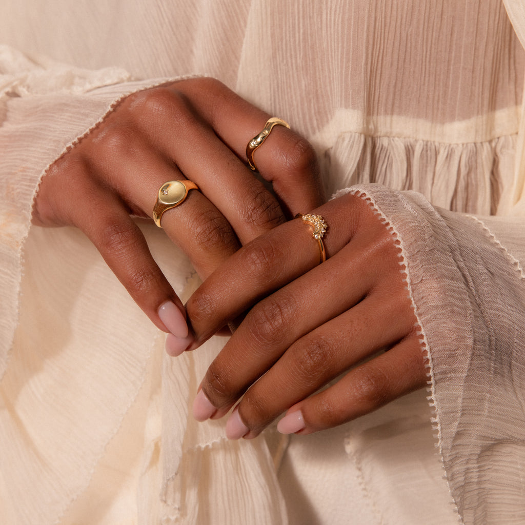 Hands with light pink nails wear three gold rings, including the Wavy Diamond Celestial Ring, resting against cream, flowy textured fabric.