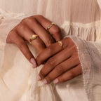 Hands with light pink nails wear three gold rings, including the Wavy Diamond Celestial Ring, resting against cream, flowy textured fabric.