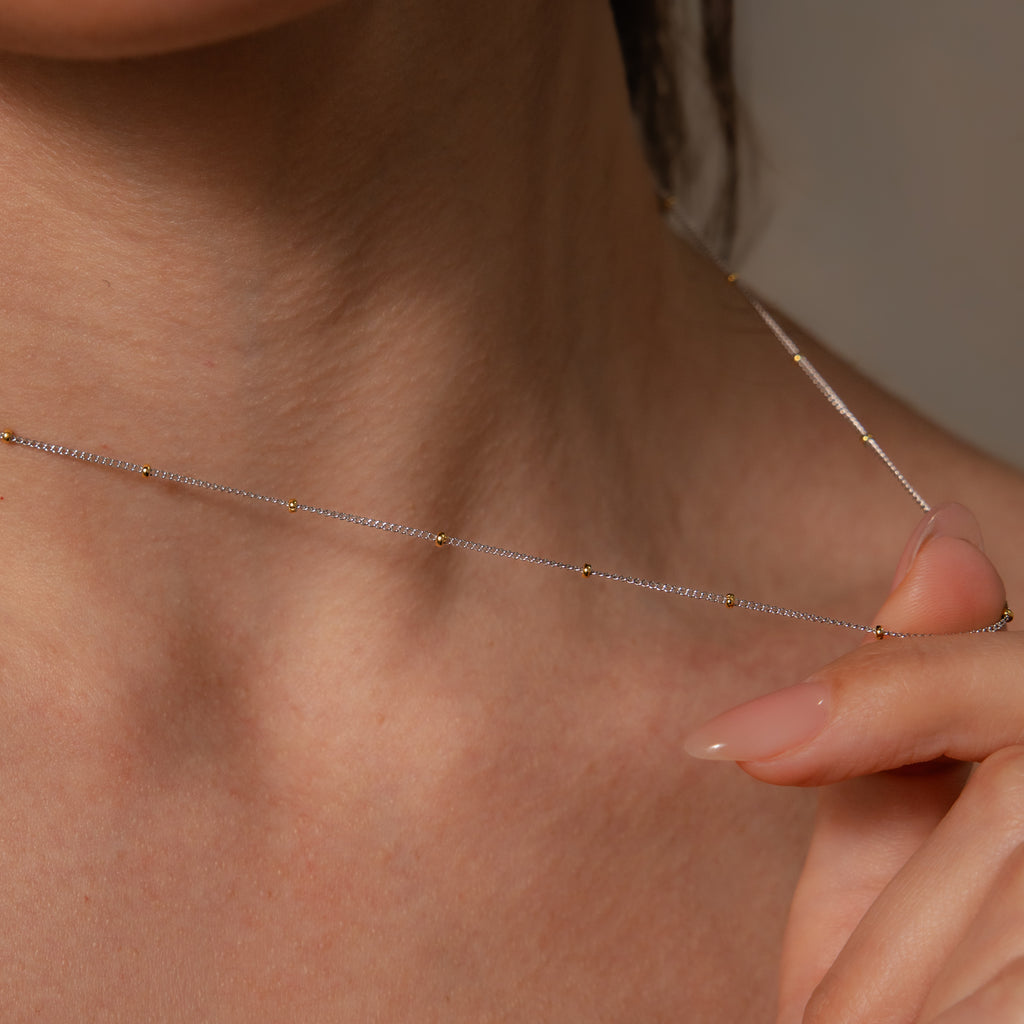 A close-up of a woman’s neck and hand showcasing the Mixed Metal Beaded Necklace featuring small gold beads, perfect for layering.