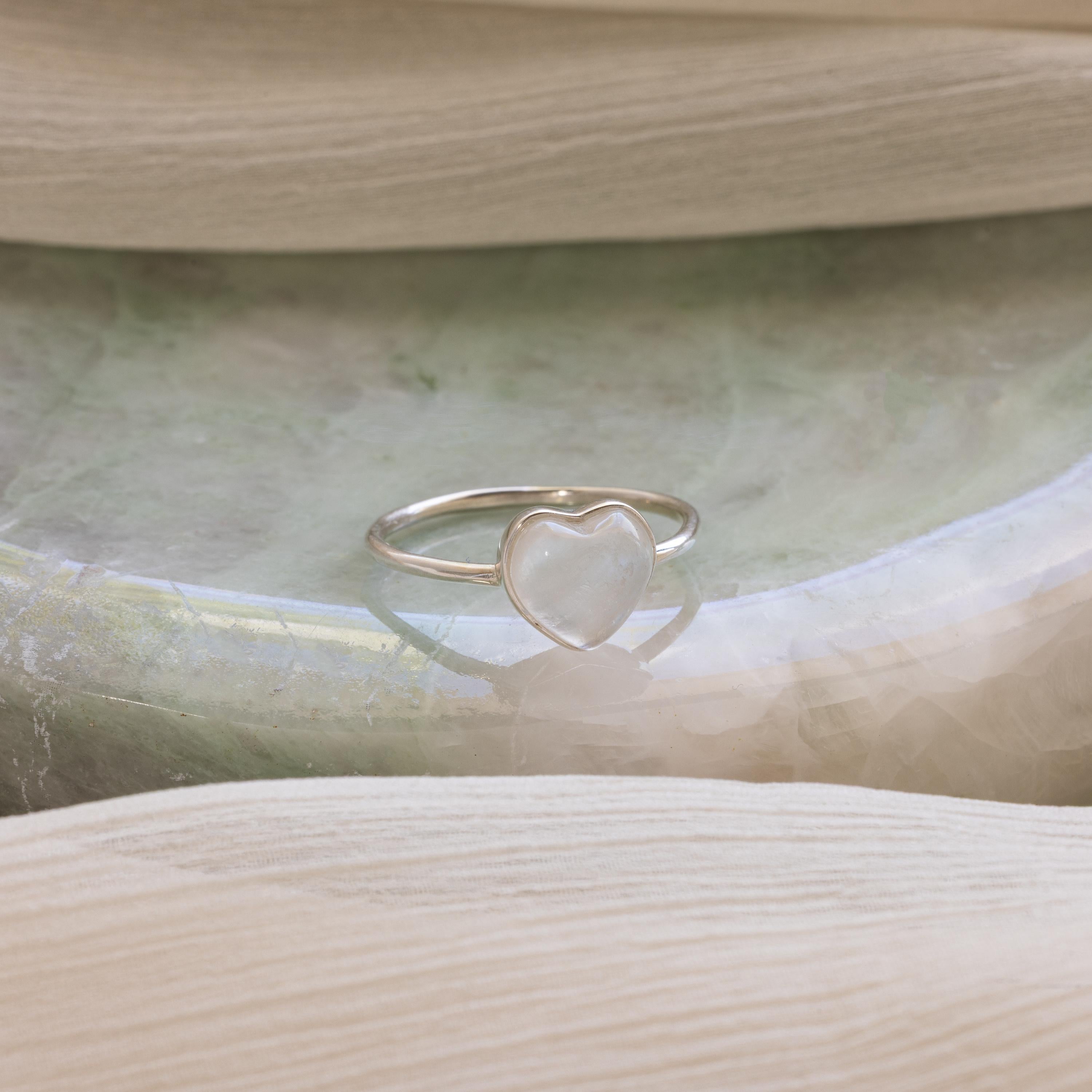 The Moonstone Heart Ring is elegantly displayed on a soft green and beige marbled surface, highlighting its luminous heart-shaped stone.