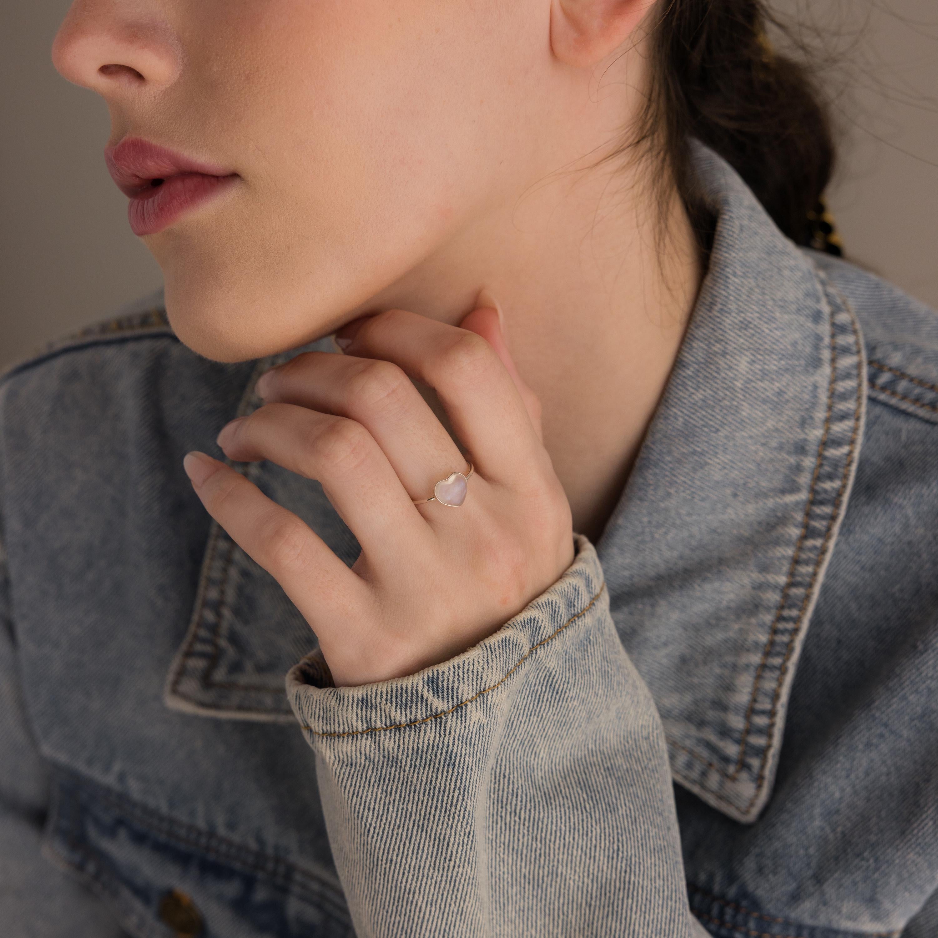 A woman wearing a denim jacket showcases the Moonstone Heart Ring, its luminous heart-shaped stone reflecting light as she rests her hand near her chin.
