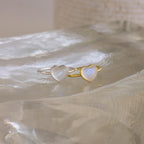 A silver and a gold heart-shaped ring, inspired by the Moonstone Heart Ring, are displayed on a clear, textured surface—each radiating elegance.