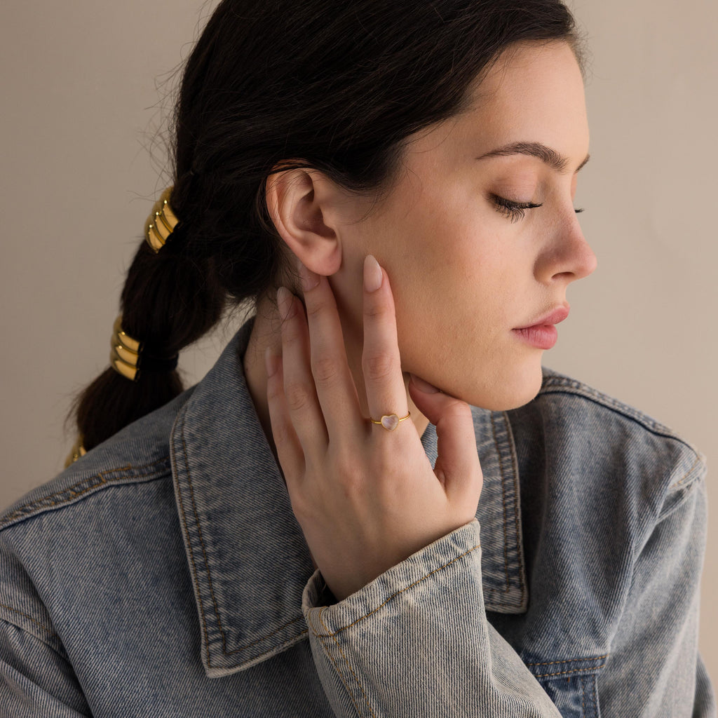 A woman in a denim jacket touches her face, showing off the Moonstone Heart Ring and braided hair with gold accessories.