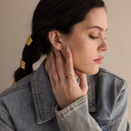 A woman in a denim jacket touches her face, showing off the Moonstone Heart Ring and braided hair with gold accessories.