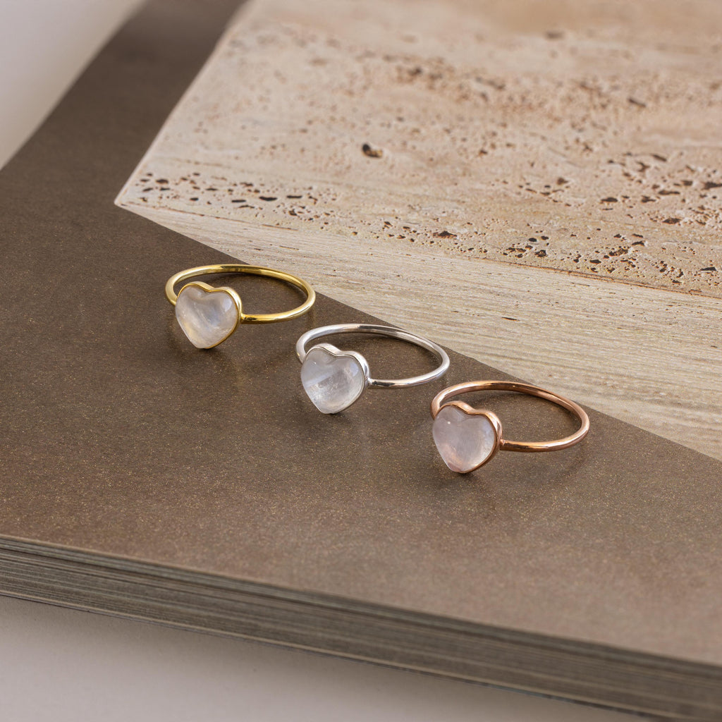 Three Moonstone Heart Rings in gold, silver, and rose gold bands—each featuring a luminous heart-shaped moonstone—are beautifully displayed on an open book.
