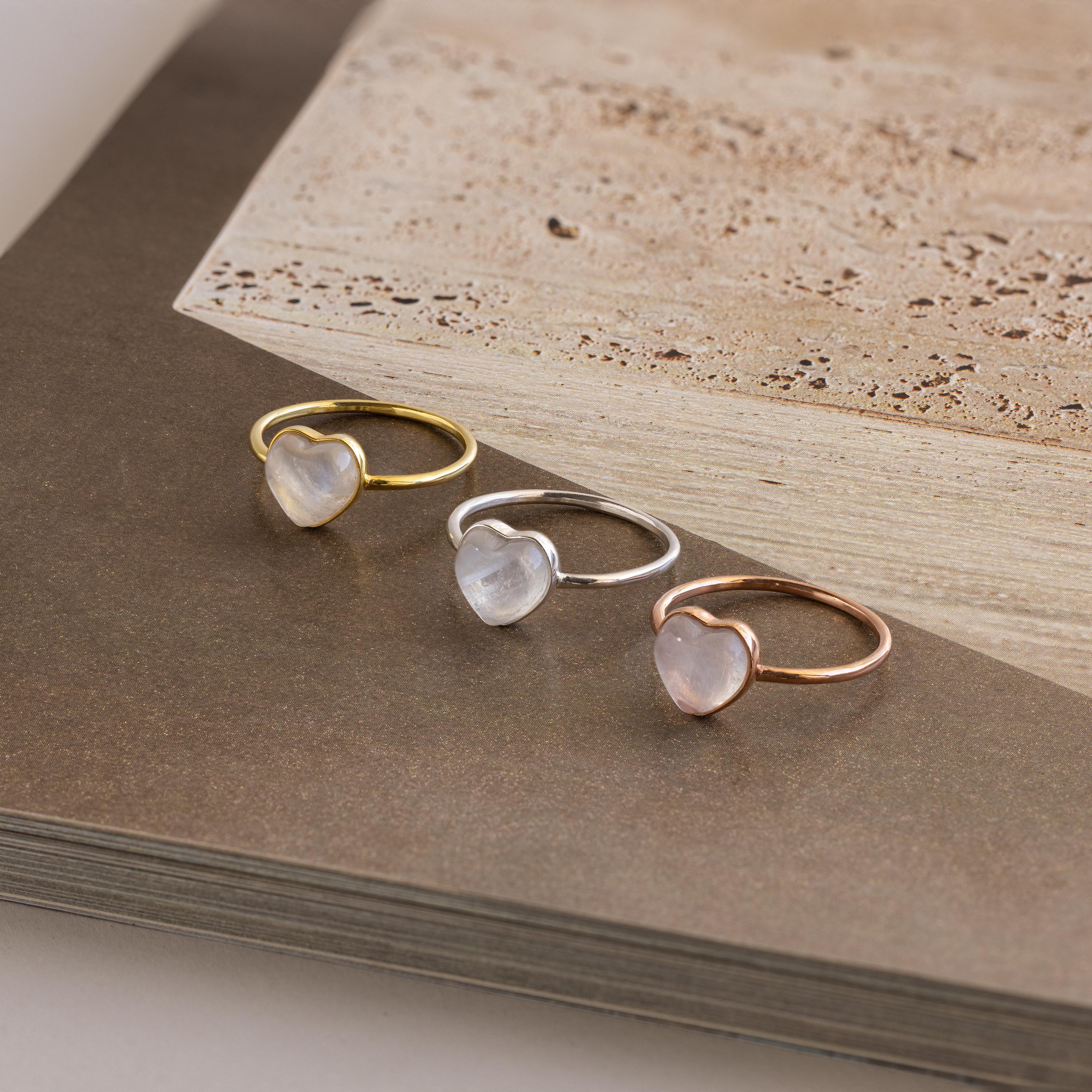 Three Moonstone Heart Rings in gold, silver, and rose gold bands—each featuring a luminous heart-shaped moonstone—are beautifully displayed on an open book.