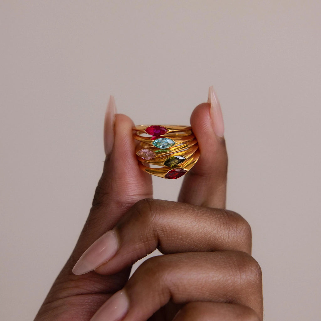 A hand with long nude nails holds stacked gold rings, including the Marquise Birthstone Signet Ring with a marquise-cut gemstone and vibrant birthstones, set against a neutral background for an elegant statement look.