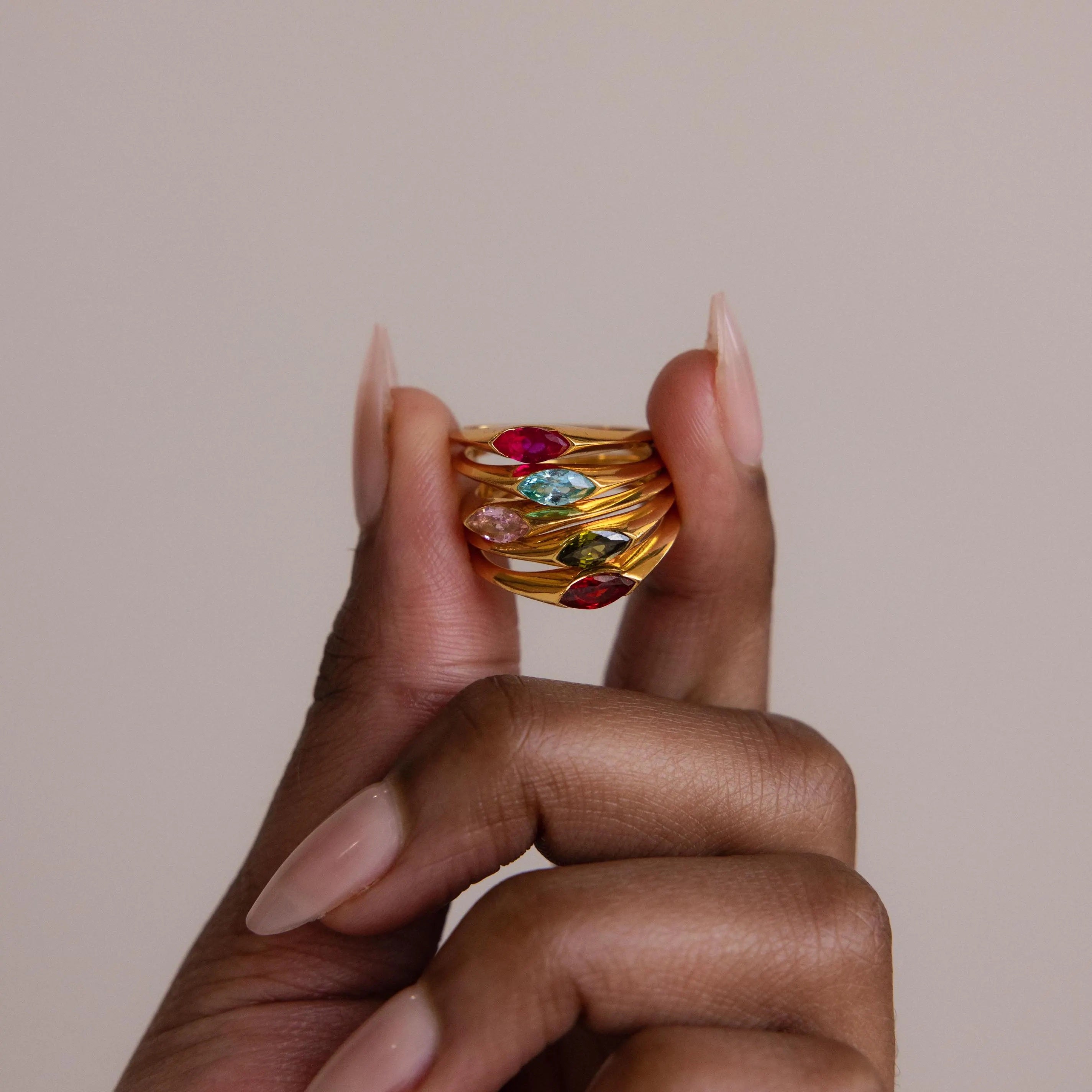 A hand with long nude nails holds stacked gold rings, including the Marquise Birthstone Signet Ring with a marquise-cut gemstone and vibrant birthstones, set against a neutral background for an elegant statement look.