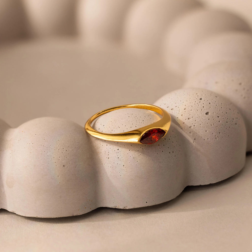 The Marquise Birthstone Signet Ring, featuring a marquise-cut red gemstone, is displayed elegantly on a textured, rounded beige surface.