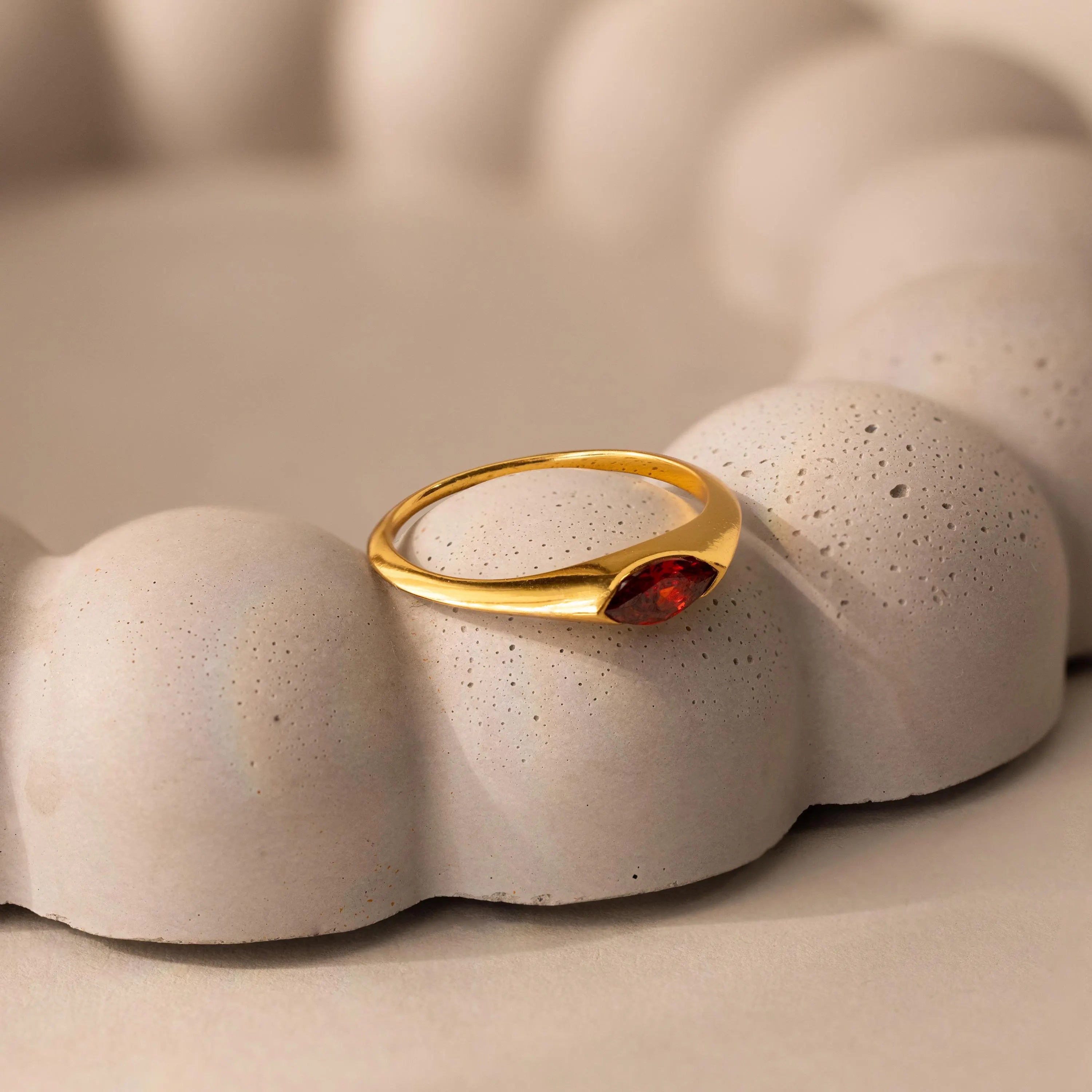The Marquise Birthstone Signet Ring, featuring a marquise-cut red gemstone, is displayed elegantly on a textured, rounded beige surface.