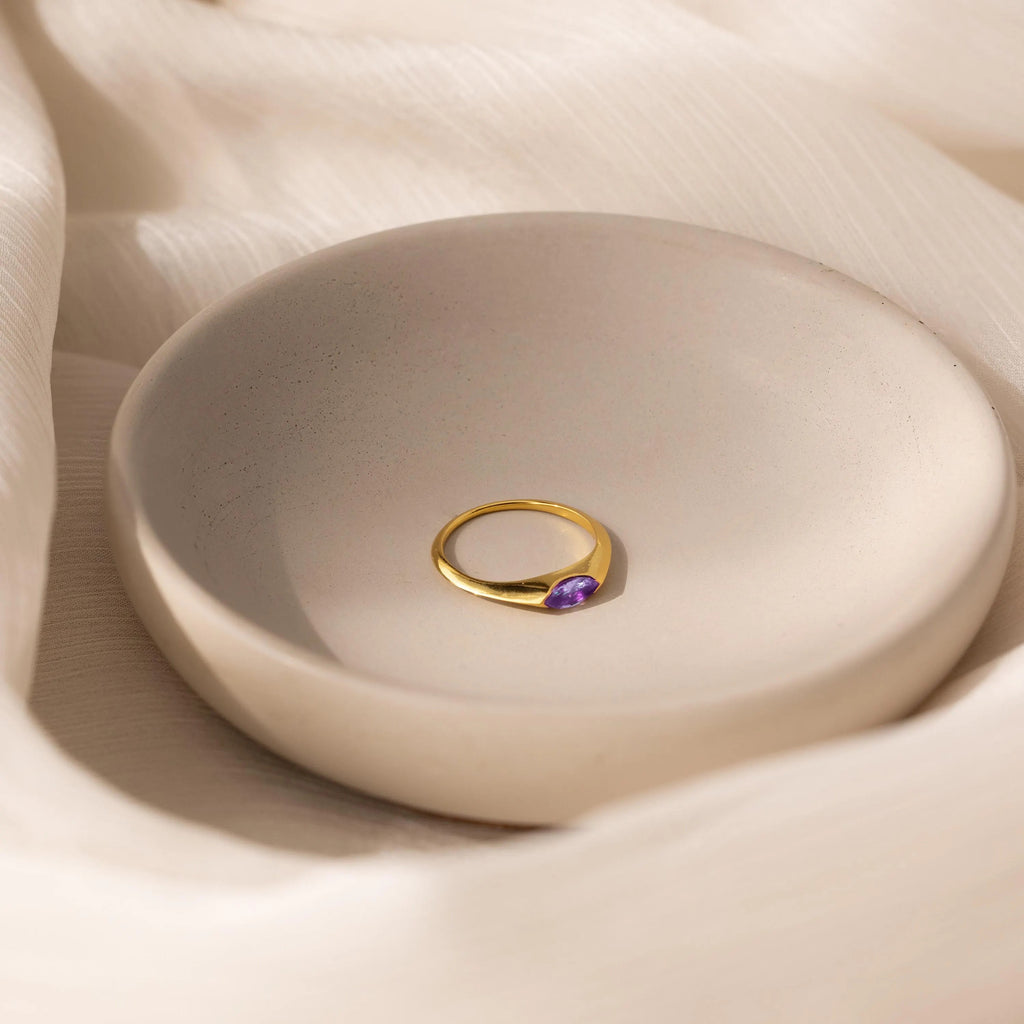 The Marquise Birthstone Signet Ring, featuring a purple gemstone, is displayed in a simple round cream-colored dish atop soft fabric.