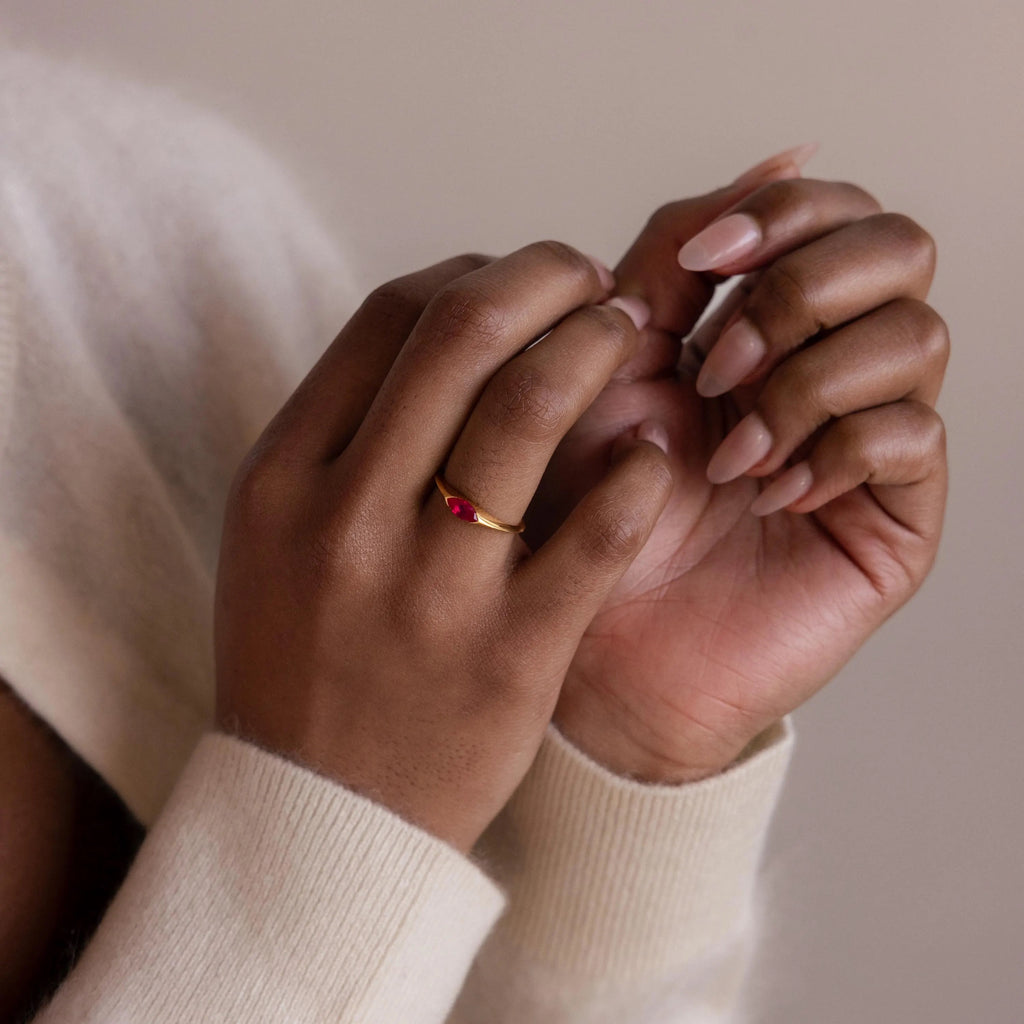 Hands with nude nails wearing the Marquise Birthstone Signet Ring, featuring a red marquise-cut gemstone, paired with a cream sweater against a neutral background.