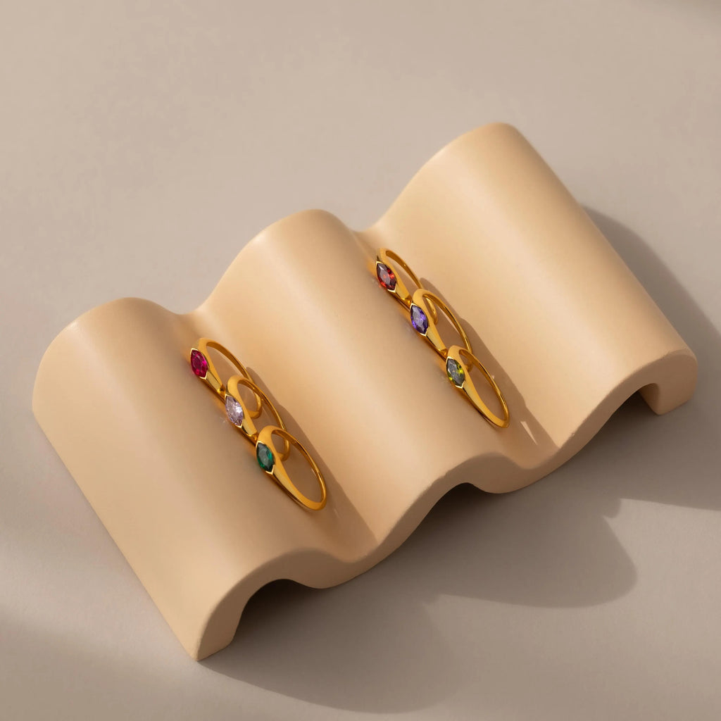 Three gold rings, including the Marquise Birthstone Signet Ring, are displayed on a wavy beige jewelry holder set against a neutral background.