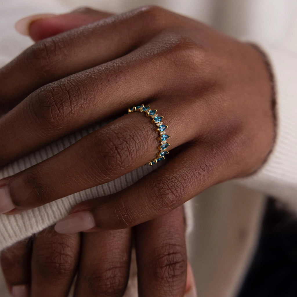 A hand wearing the Marquise Infinity Birthstone Ring, featuring a gold infinity design with small blue gemstones, rests gently on another hand.