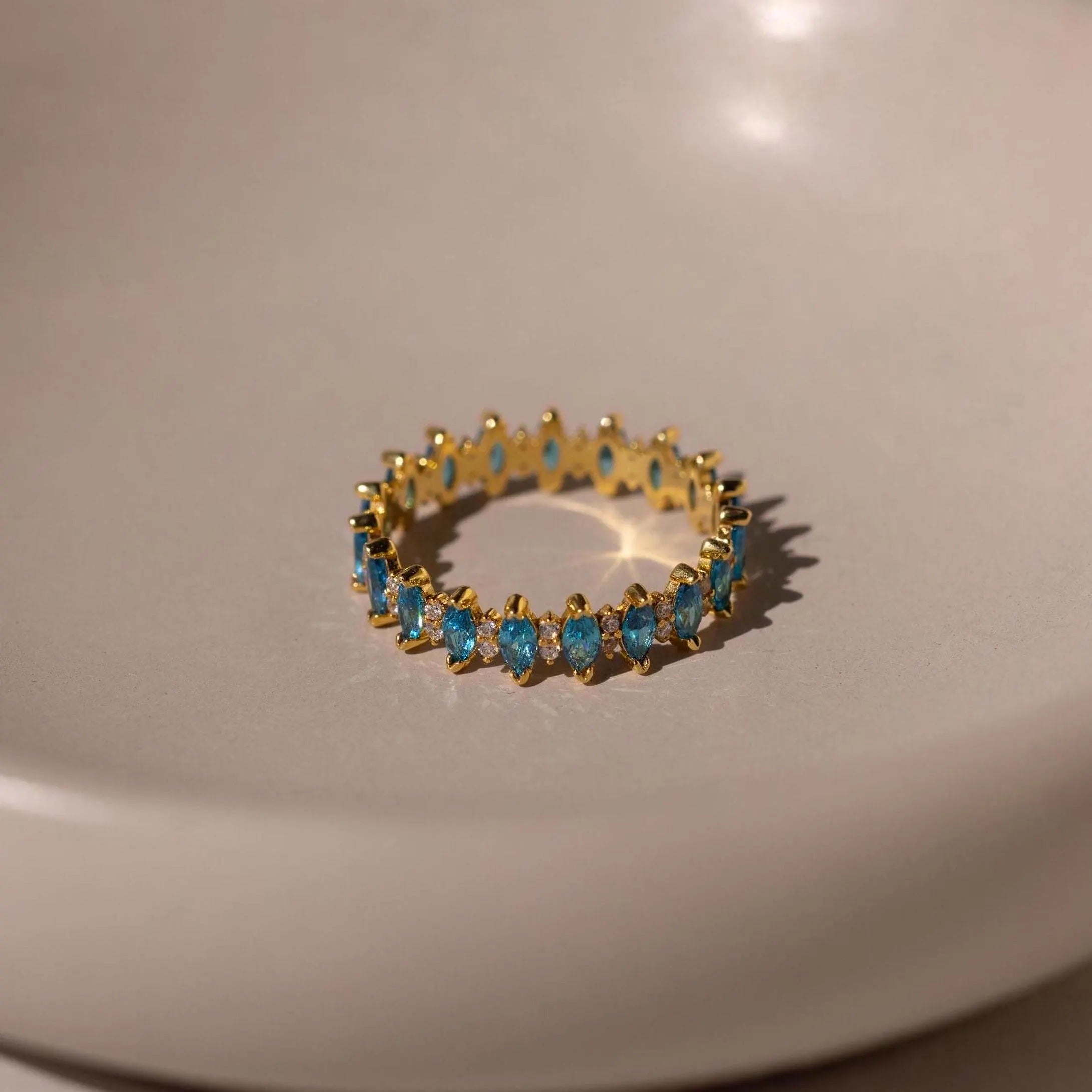 The Marquise Infinity Birthstone Ring features a continuous row of small blue gemstones in an elegant infinity design, displayed on a smooth, light-colored surface—the perfect personalized ring.