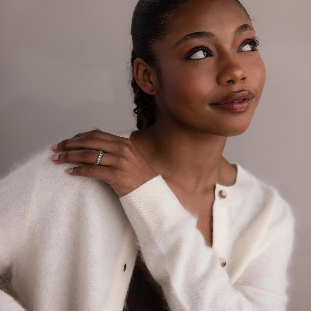 A woman in a white sweater smiles softly, hand on shoulder, wearing the Marquise Infinity Birthstone Ring featuring its unique infinity design.