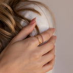 A delicate hand with neutral manicured nails wears a plain gold band paired with a matching amethyst-stone ring