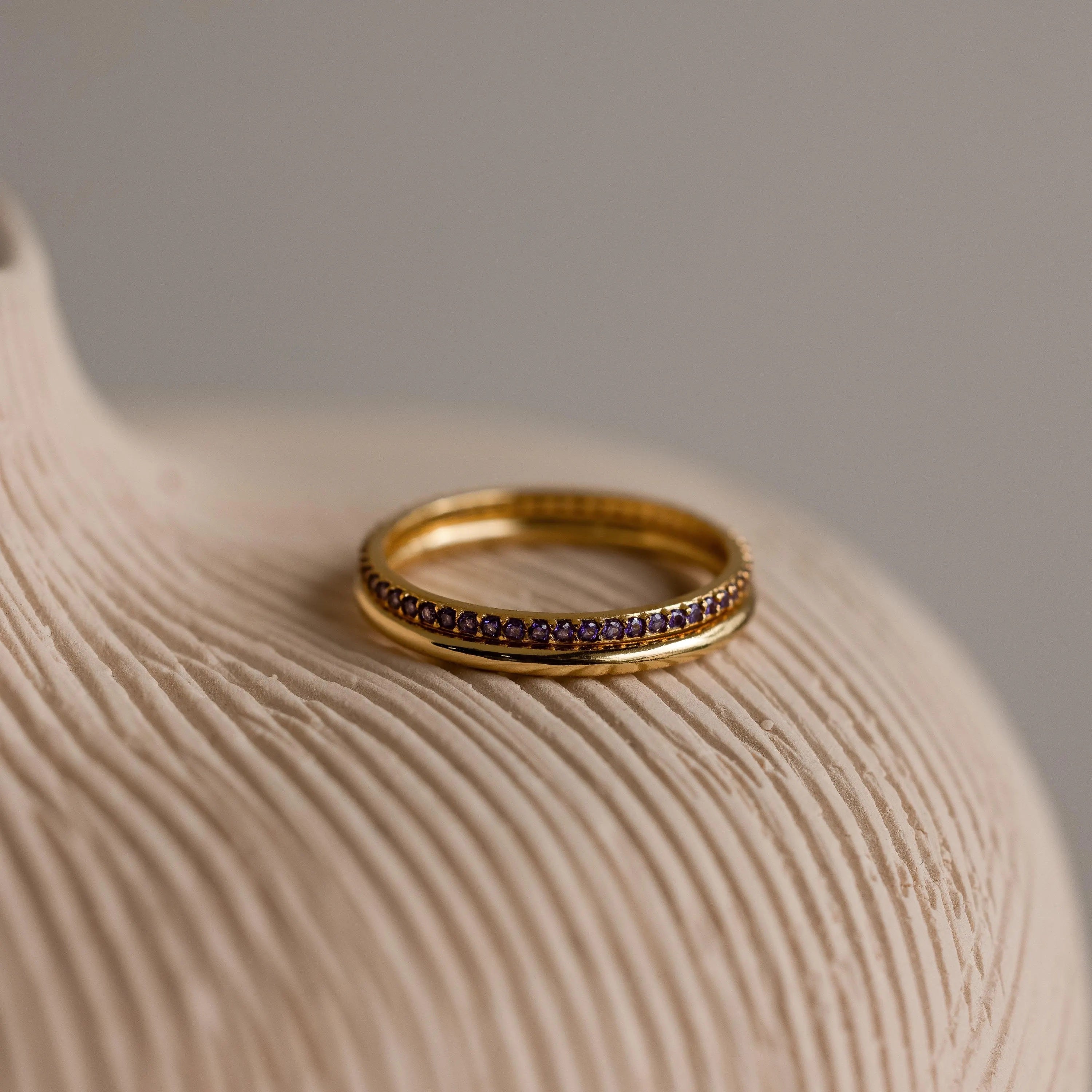 A gold band ring and an 18K gold tennis ring with purple gemstones, placed on a neutral surface.
