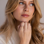 A portrait view of a woman resting her chin on her hand, displaying a neatly stacked pair of gold rings with purple stones and a plain band.