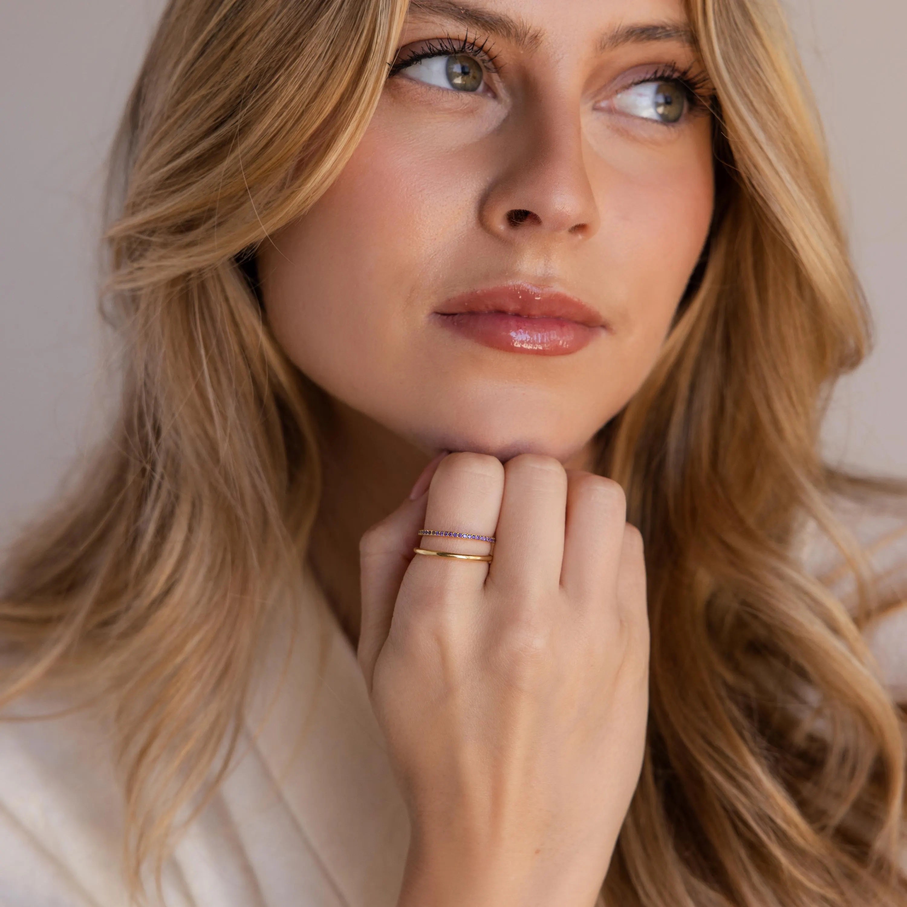 A portrait view of a woman resting her chin on her hand, displaying a neatly stacked pair of gold rings with purple stones and a plain band.