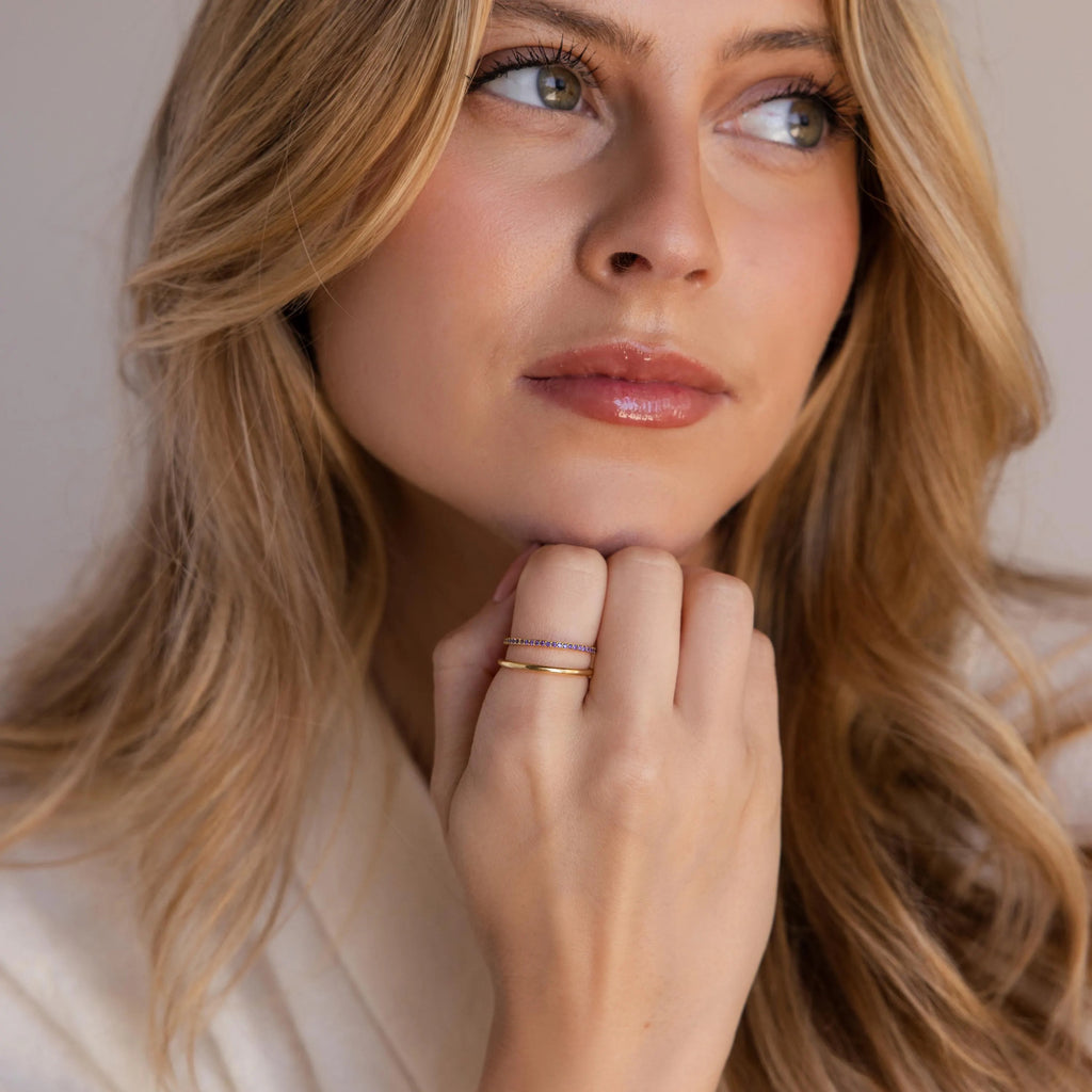 A portrait view of a woman resting her chin on her hand, displaying a neatly stacked pair of gold rings with purple stones and a plain band.