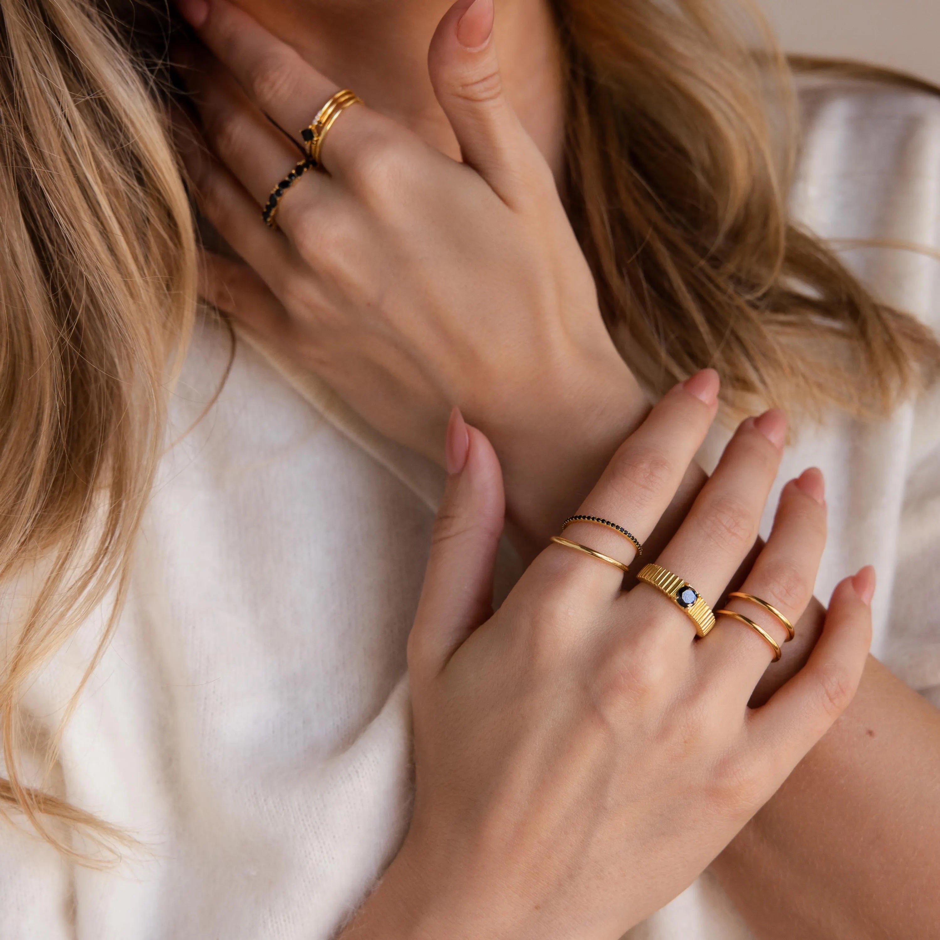 Hands adorned with a mix of slim and textured gold rings, including multiple birthstone bands in black. 