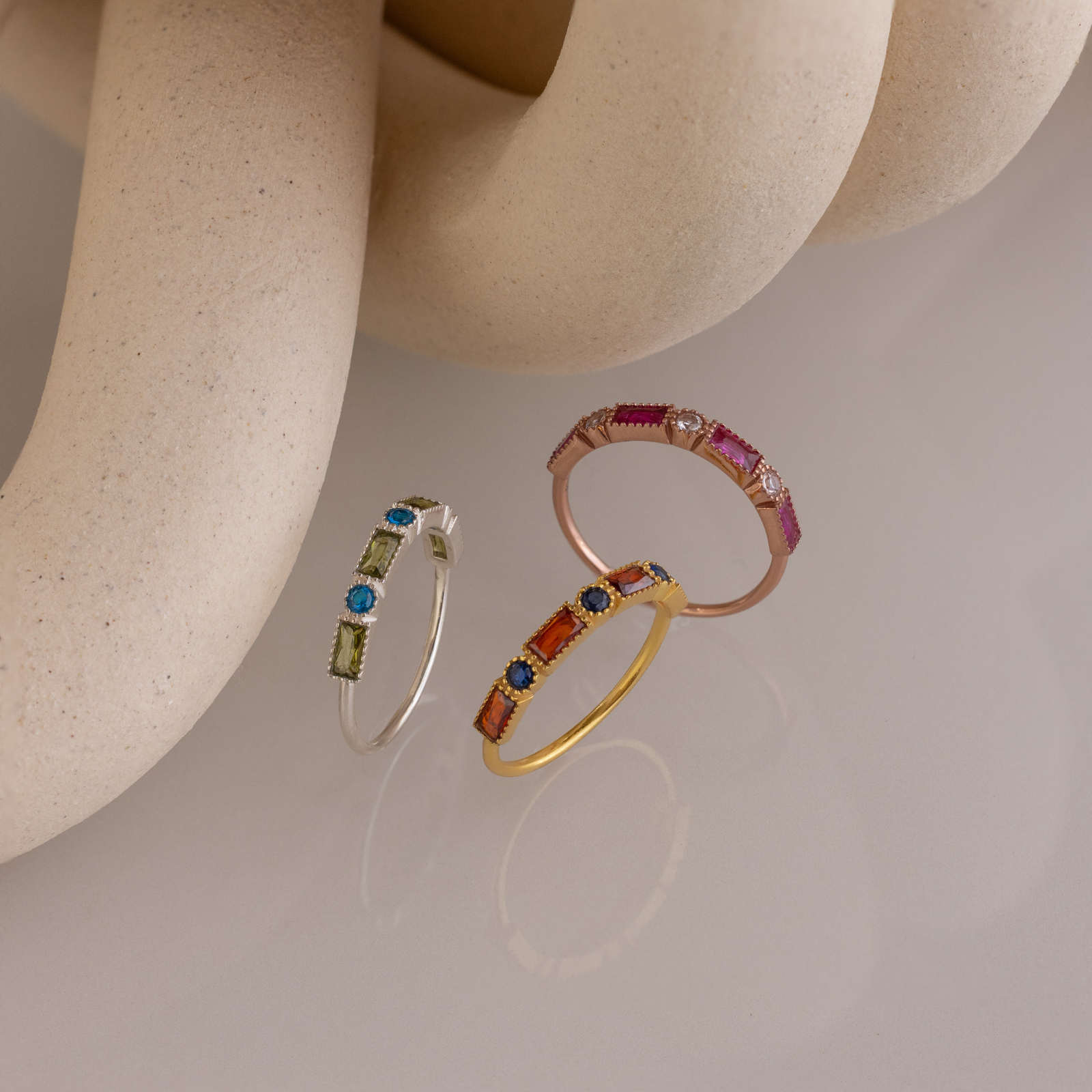 The Mixed Art Deco Birthstone Ring, along with two other colorful gemstone rings, is displayed on a smooth, light surface with abstract beige shapes in the background.