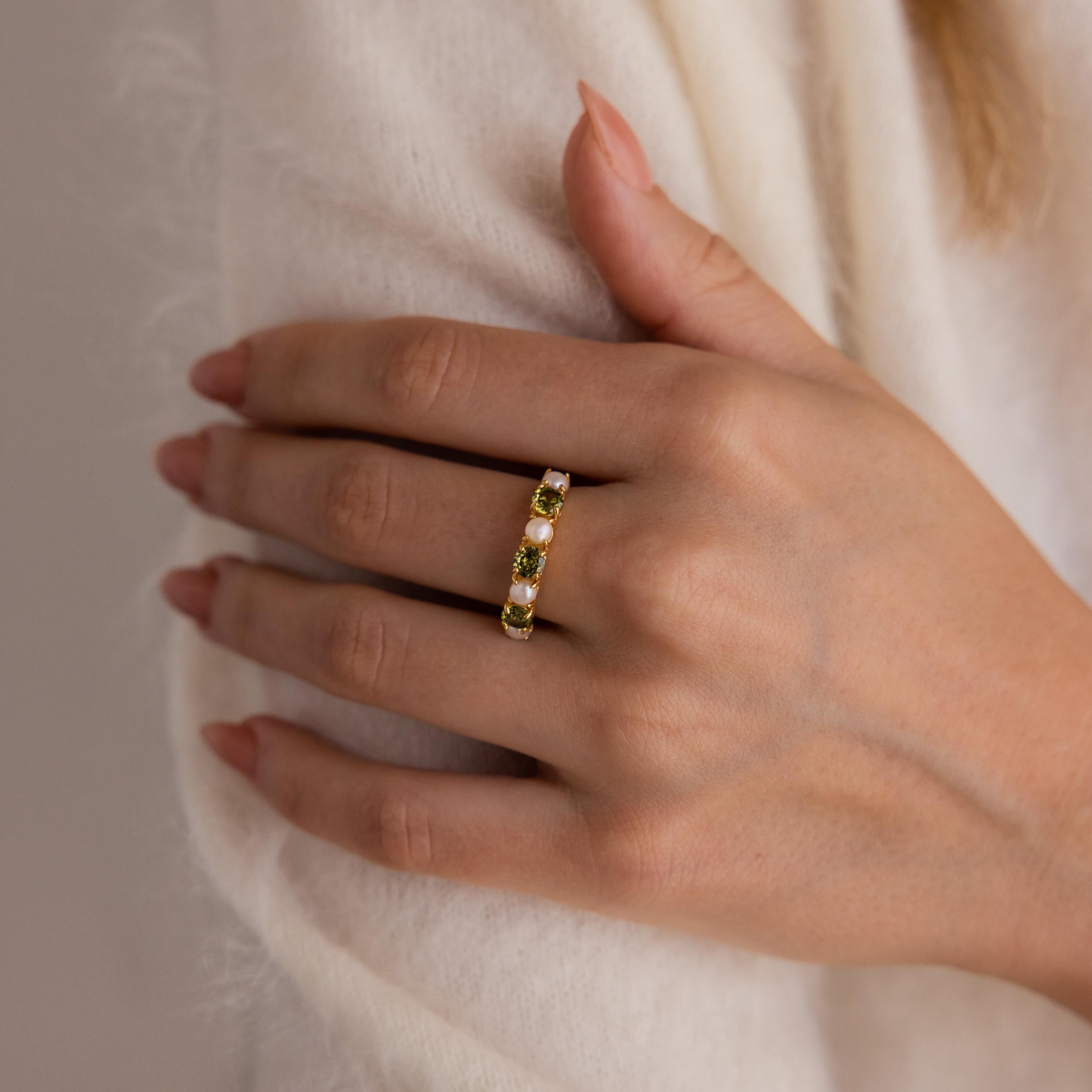 A hand with neatly manicured nails rests on cream-colored fabric, wearing the Peridot & Pearl Lace Ring, a gold band adorned with three green gemstones.