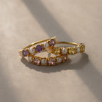 The Amethyst & Pearl Lace Ring features gold bands with alternating white, purple, and yellow stones, arranged on a beige surface in soft light—a perfect choice for subtle elegance.