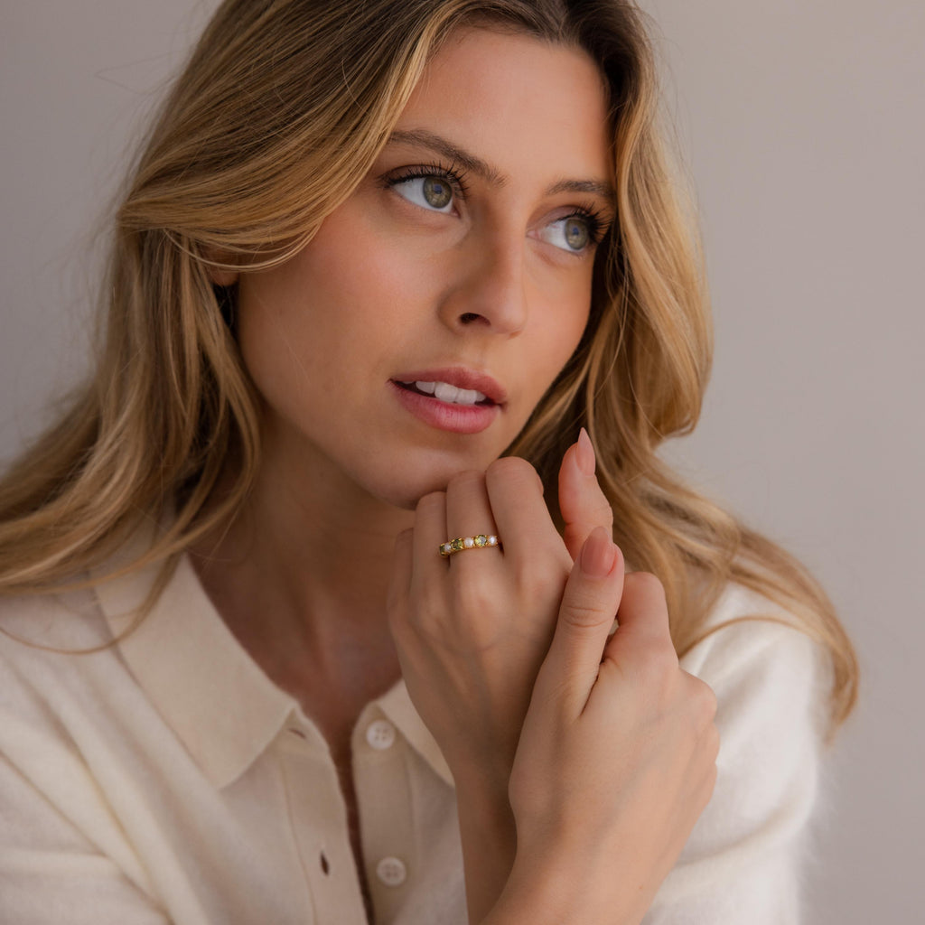 A woman with long blonde hair in a cream sweater looks to the side, displaying the Amethyst & Pearl Lace Ring—a delicate gold band on her finger.