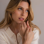 A woman with long blonde hair in a cream sweater looks to the side, displaying the Amethyst & Pearl Lace Ring—a delicate gold band on her finger.