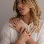 A woman with blonde hair wearing a cream top showcases several gold rings, including the Peridot & Pearl Lace Ring, on her hands as she looks to the side against a neutral background.