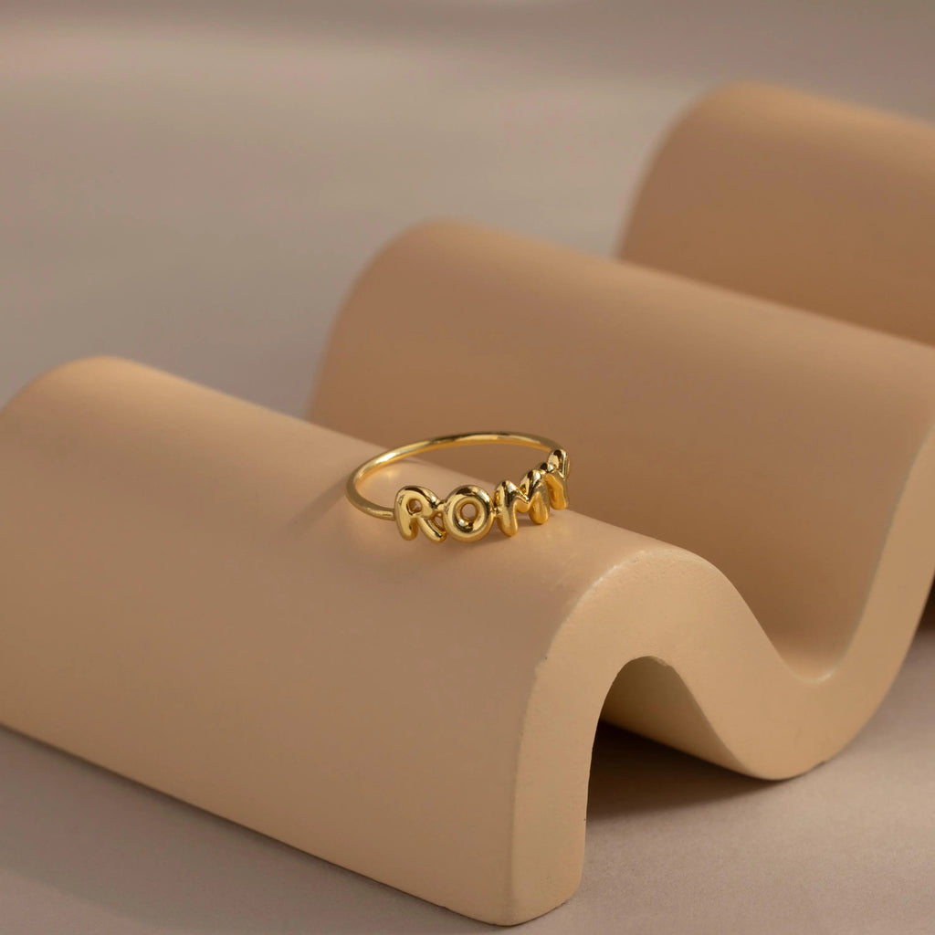 Gold bubble letter ring with the name "ROMY' resting on a neutral-tone wavy ceramic dish.