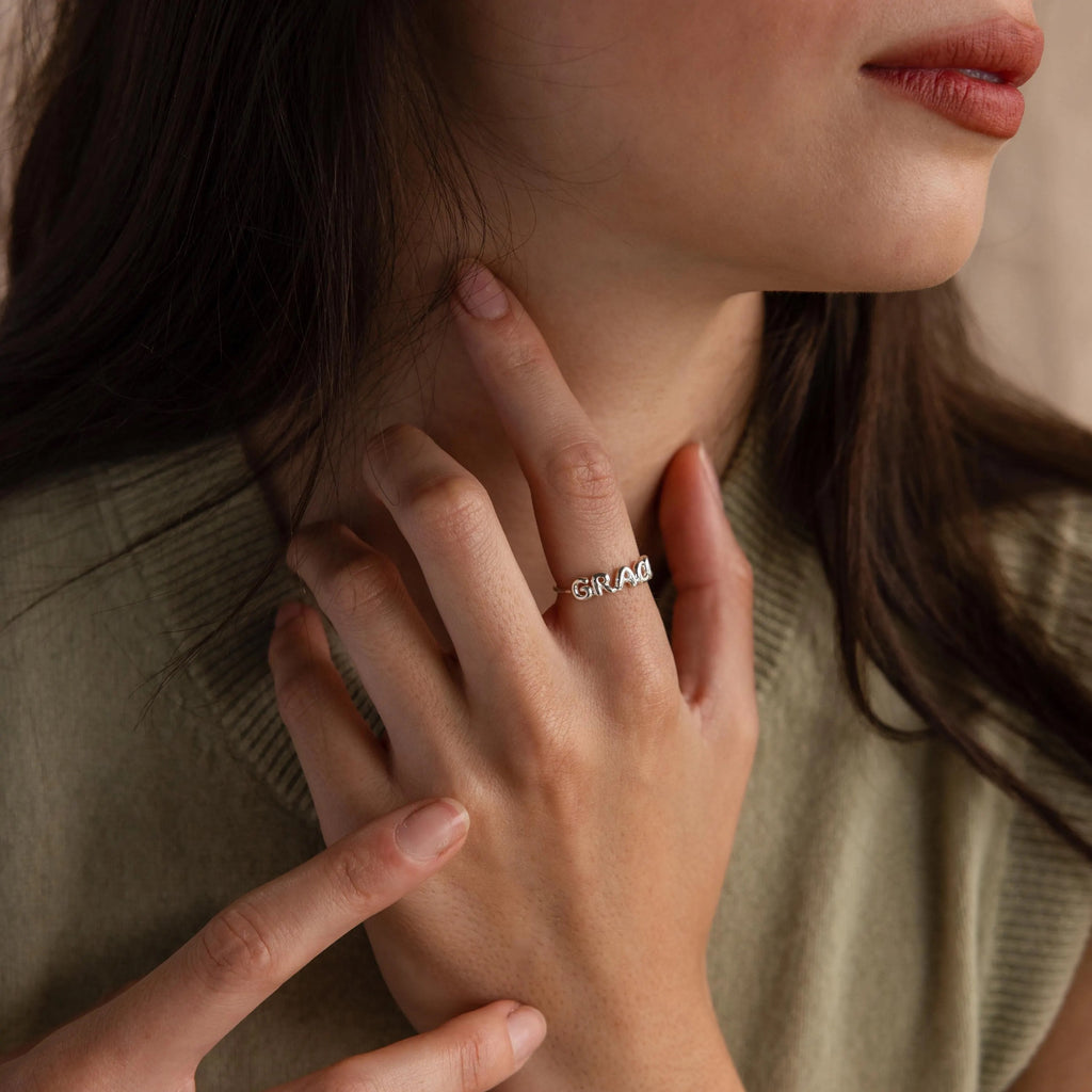 A woman touches her neck, showcasing the Bubble Name Ring with engraved 3D letters on her finger.