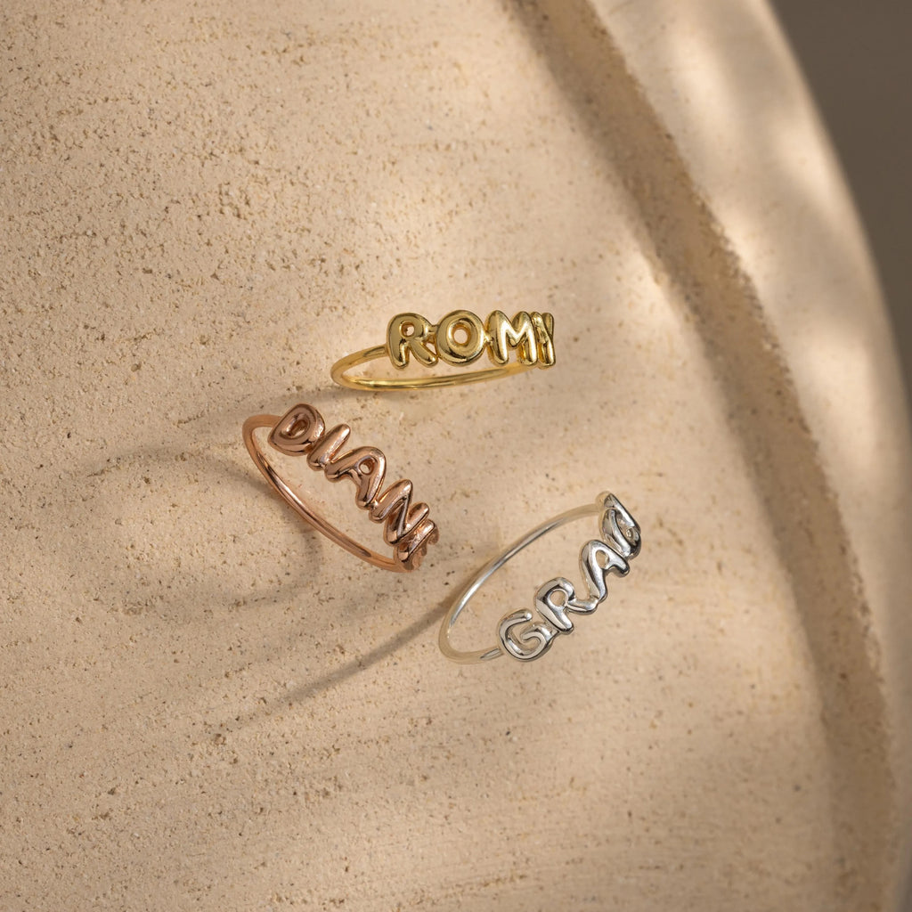 Three Bubble Name Rings with 3D letters spelling ROMY, DJANI, and GRAN rest on a textured beige surface—each a unique personalized ring, ideal for mixing and stacking.