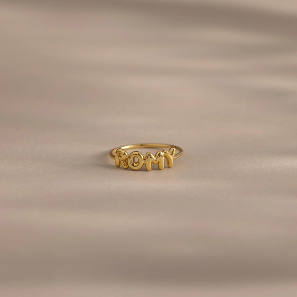 Close up of a gold bubble letter ring with the name "ROMY" resting on a neutral, beige surface.