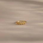Close up of a gold bubble letter ring with the name "ROMY" resting on a neutral, beige surface.
