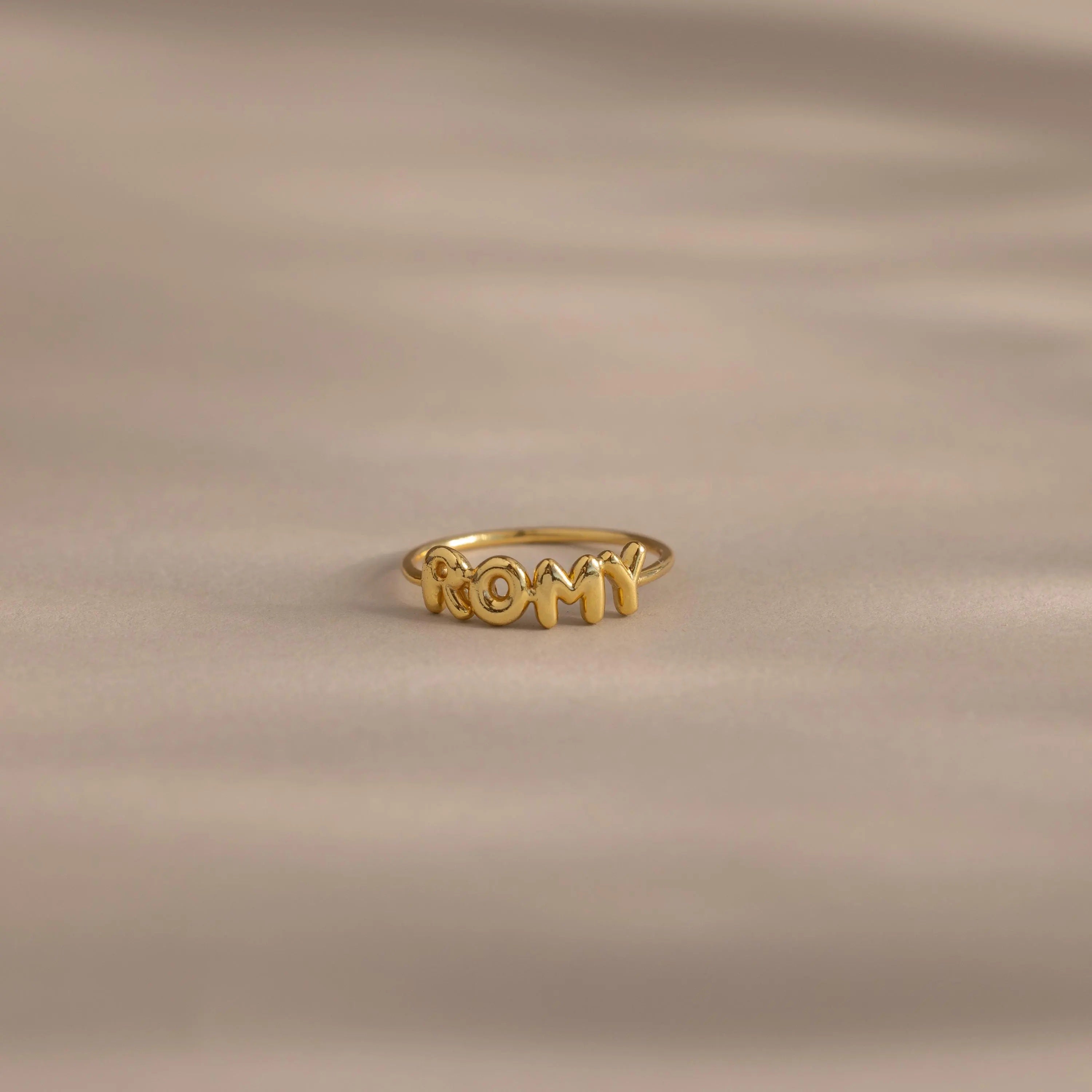 Close up of a gold bubble letter ring with the name "ROMY" resting on a neutral, beige surface.
