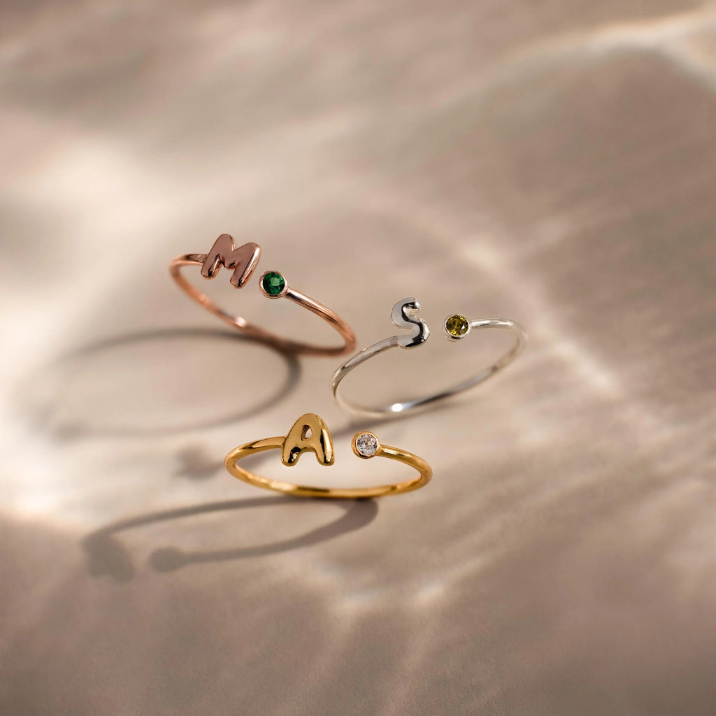 Close up of 3 dainty bubble letter gemstone rings, including a rose gold ring with the initial "H" and a green emerald birthstone, a silver ring with the initial "S" and a green peridot birthstone, and a gold ring with the initial "A" and a clear diamond birthstone.