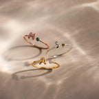 Close up of 3 dainty bubble letter gemstone rings, including a rose gold ring with the initial "H" and a green emerald birthstone, a silver ring with the initial "S" and a green peridot birthstone, and a gold ring with the initial "A" and a clear diamond birthstone.