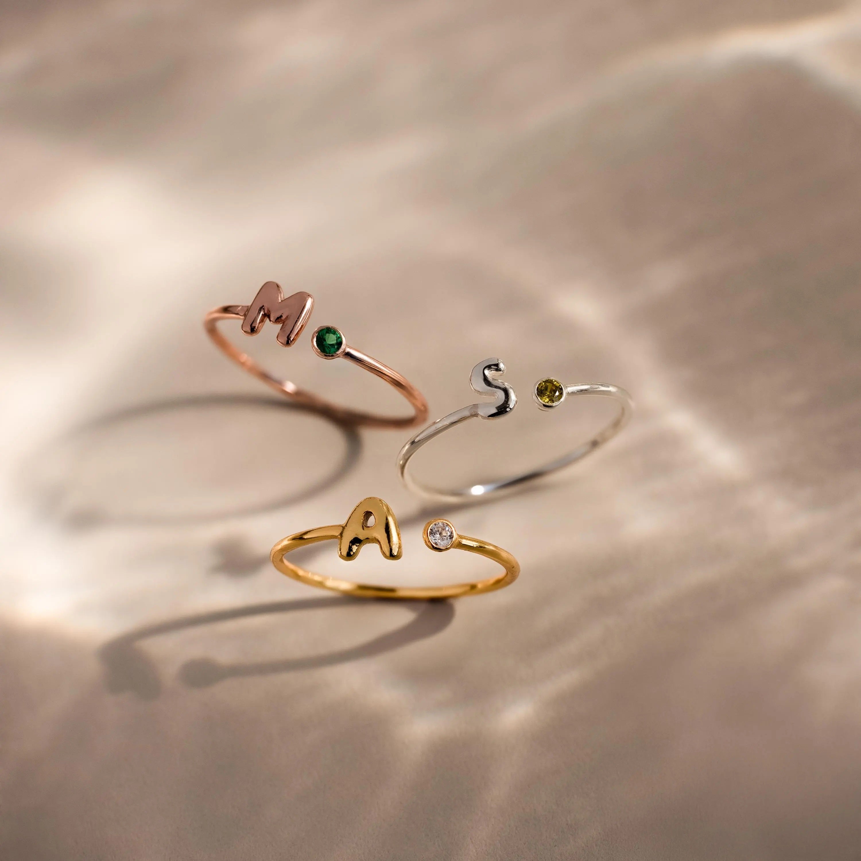 Close up of 3 dainty bubble letter gemstone rings, including a rose gold ring with the initial "H" and a green emerald birthstone, a silver ring with the initial "S" and a green peridot birthstone, and a gold ring with the initial "A" and a clear diamond birthstone.