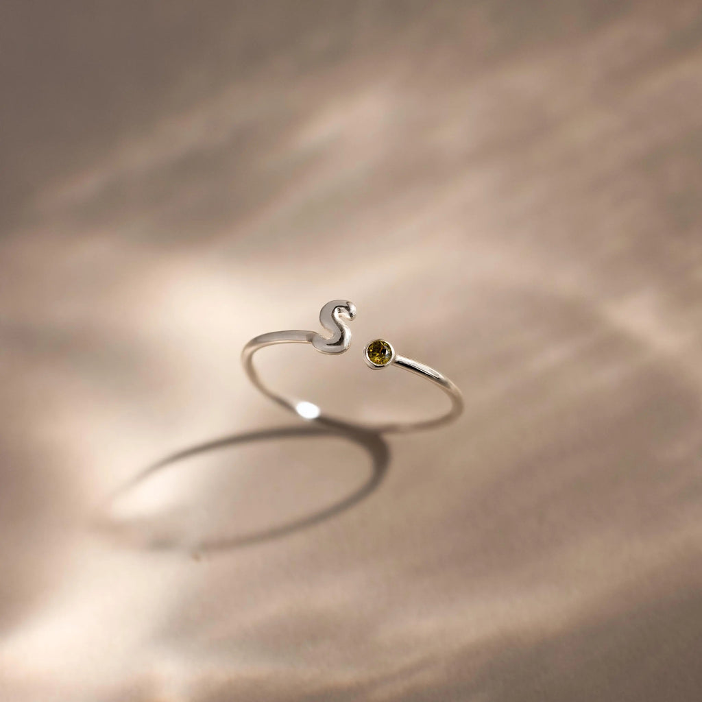 Up close of a silver bubble letter duo gemstone ring with the initial "S" and a green peridot birthstone displayed on a neutral surface.