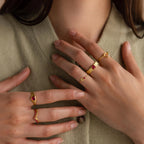Model wearing a dainty gold bubble letter birthstone duo ring with the initial "A" and a clear diamond gemstone, stacked with multiple gold rings with pink ruby gemstones.