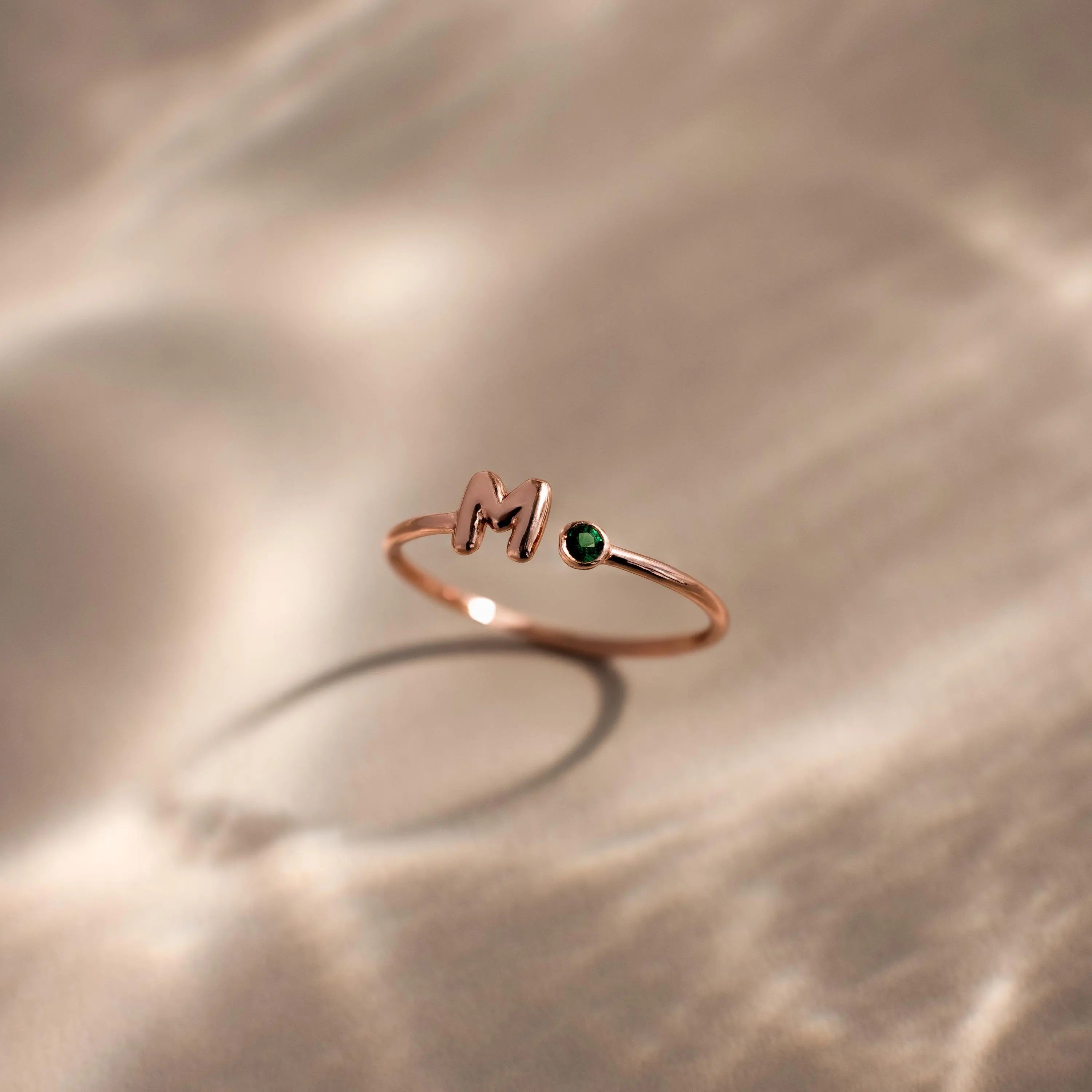 Up close of a dainty rose gold bubble letter duo gemstone ring with the initial "H" and a green emerald birthstone displayed on a neutral background.