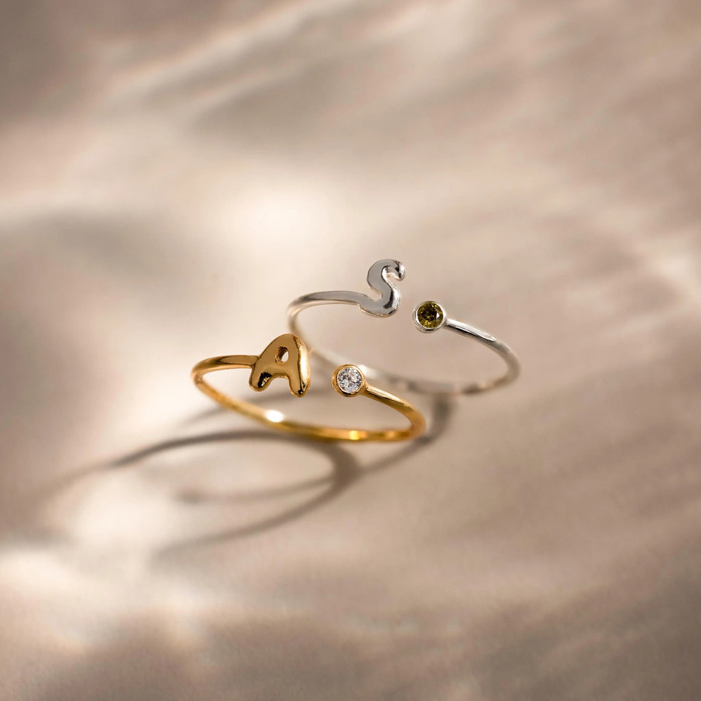 Up close of 2 bubble initial duo gemstone rings, featuring a gold ring with the letter "A" and a clear diamond birthstone and a silver ring with the letter "S" and a green peridot birthstone.