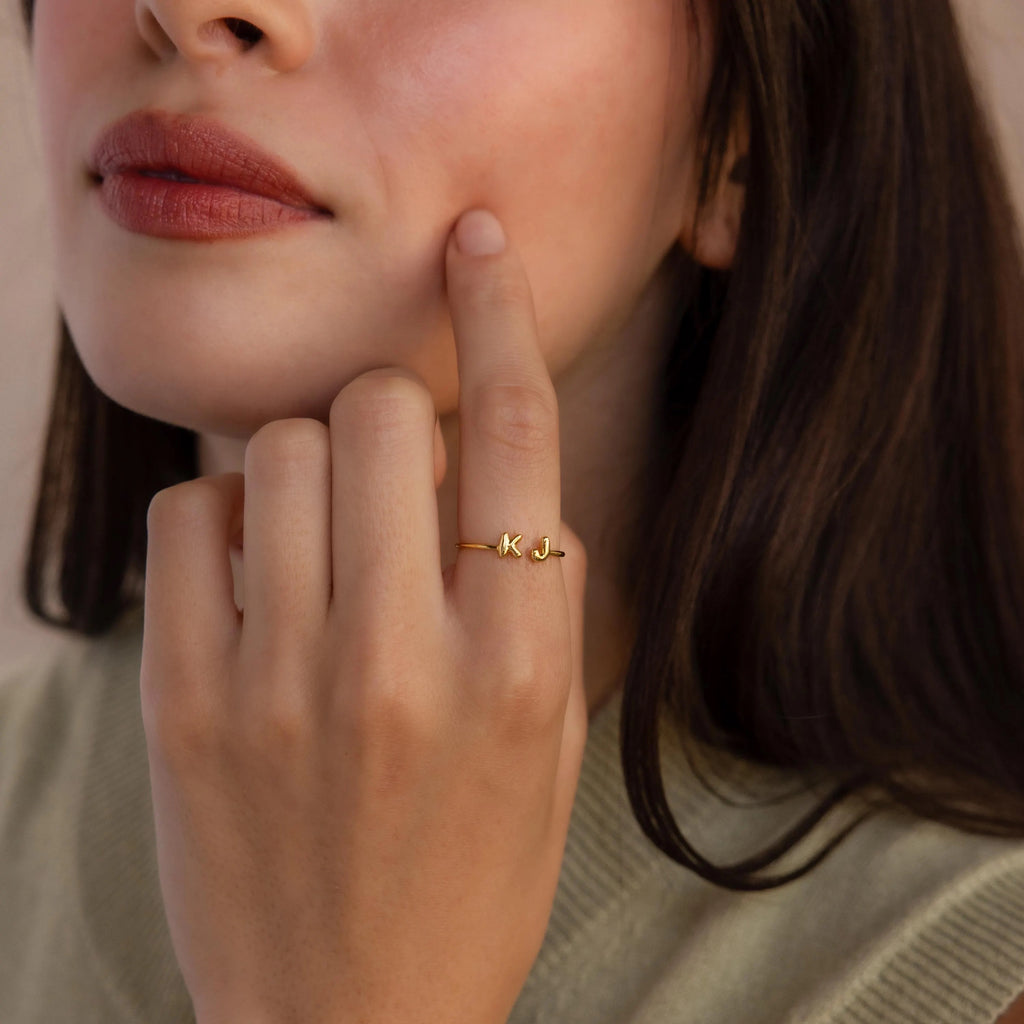 Close-up of a dainty gold double initial ring with bubble letters K and J, worn on hand with soft natural makeup and neutral clothing.