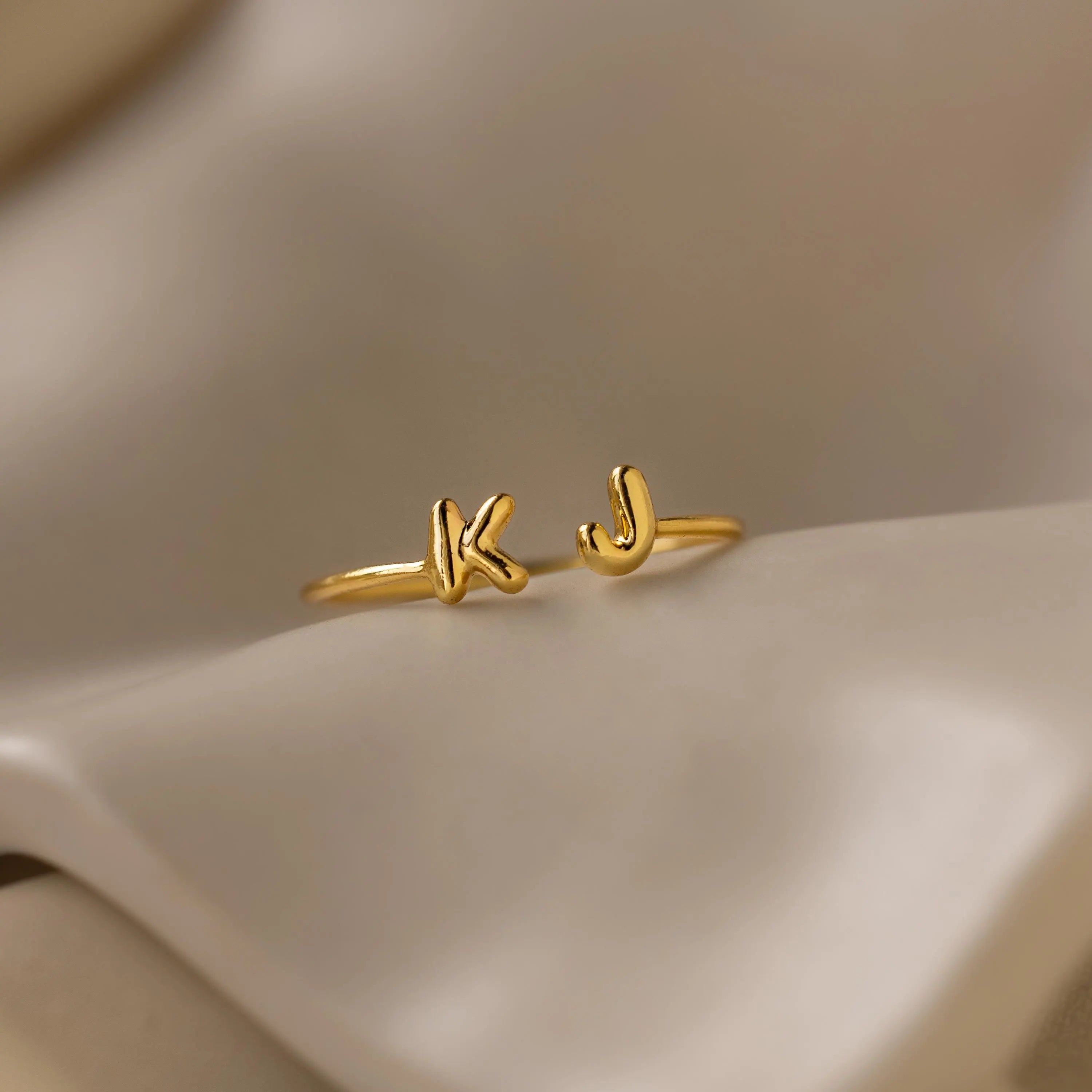 Close-up of a gold open ring featuring bubble-style initials K and J on soft satin fabric.
